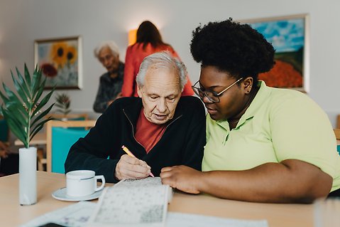 Miljö från ett äldreboende. I förgrunden en anställd sittandes med en boende vid ett bord.