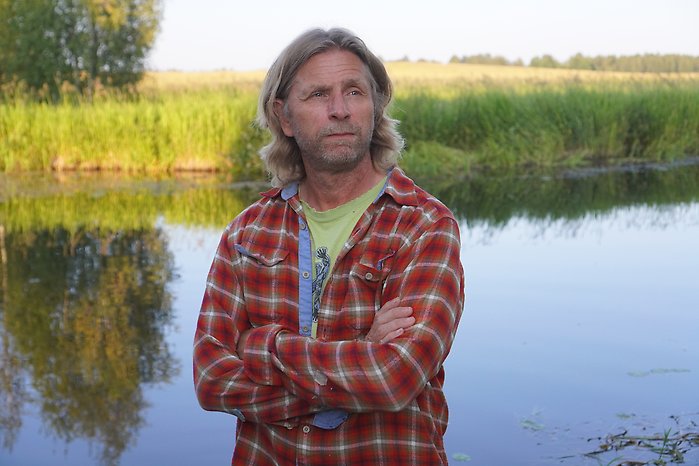 Porträttbild på konstnären Jonas. Han står med armarna i kors och tittar åt sidan. I bakgrunden syns en sjö, vass och träd.